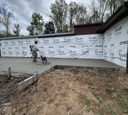 Wide view of a newly poured concrete slab in Lyles, TN beside an existing home surrounded by trees.